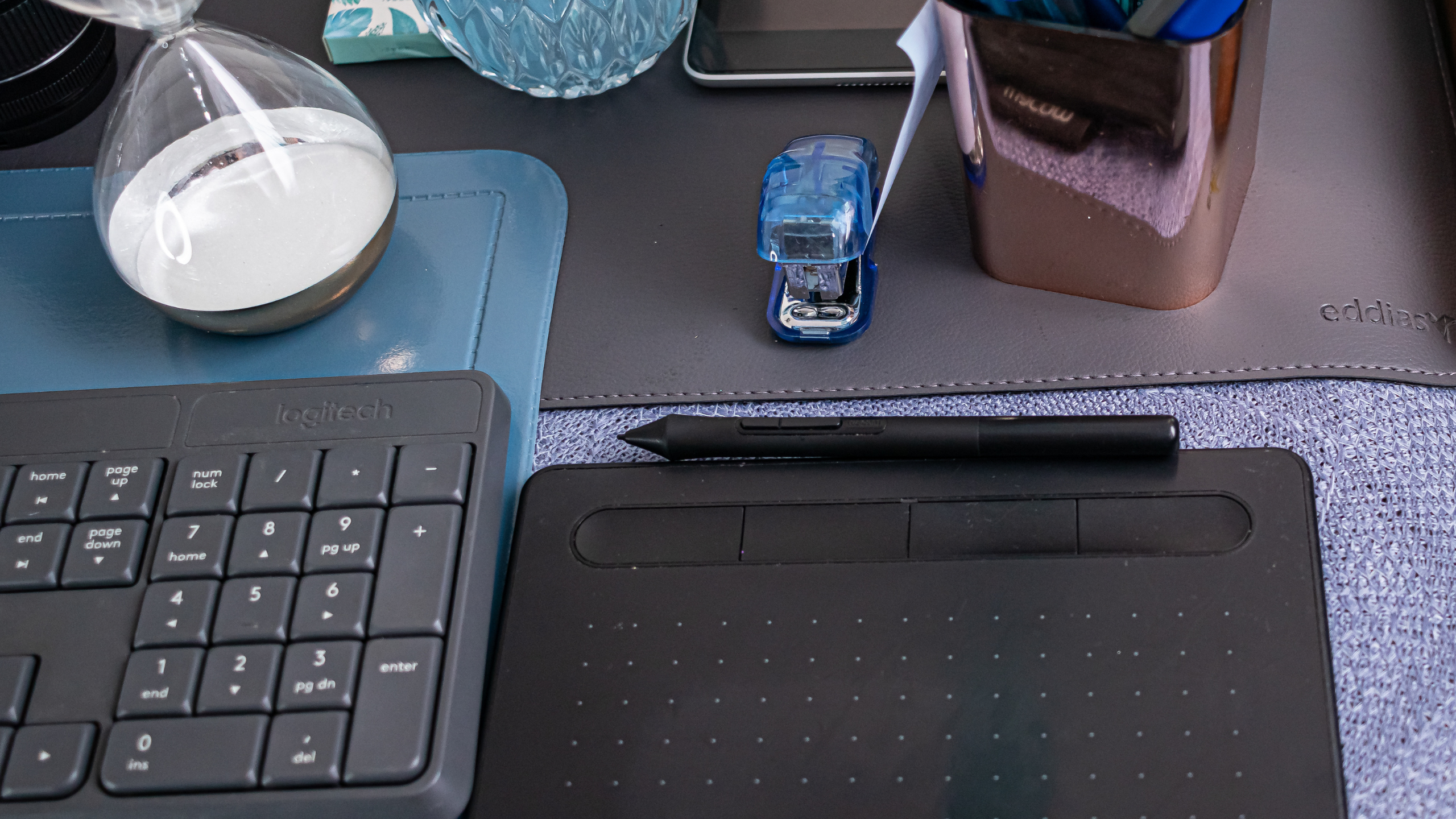 Mesa de trabalho de Carla Toscano Pós-Produção Fotográfica, com teclado Logitech, mesa digitalizadora, ampulheta e itens de escritório organizados.