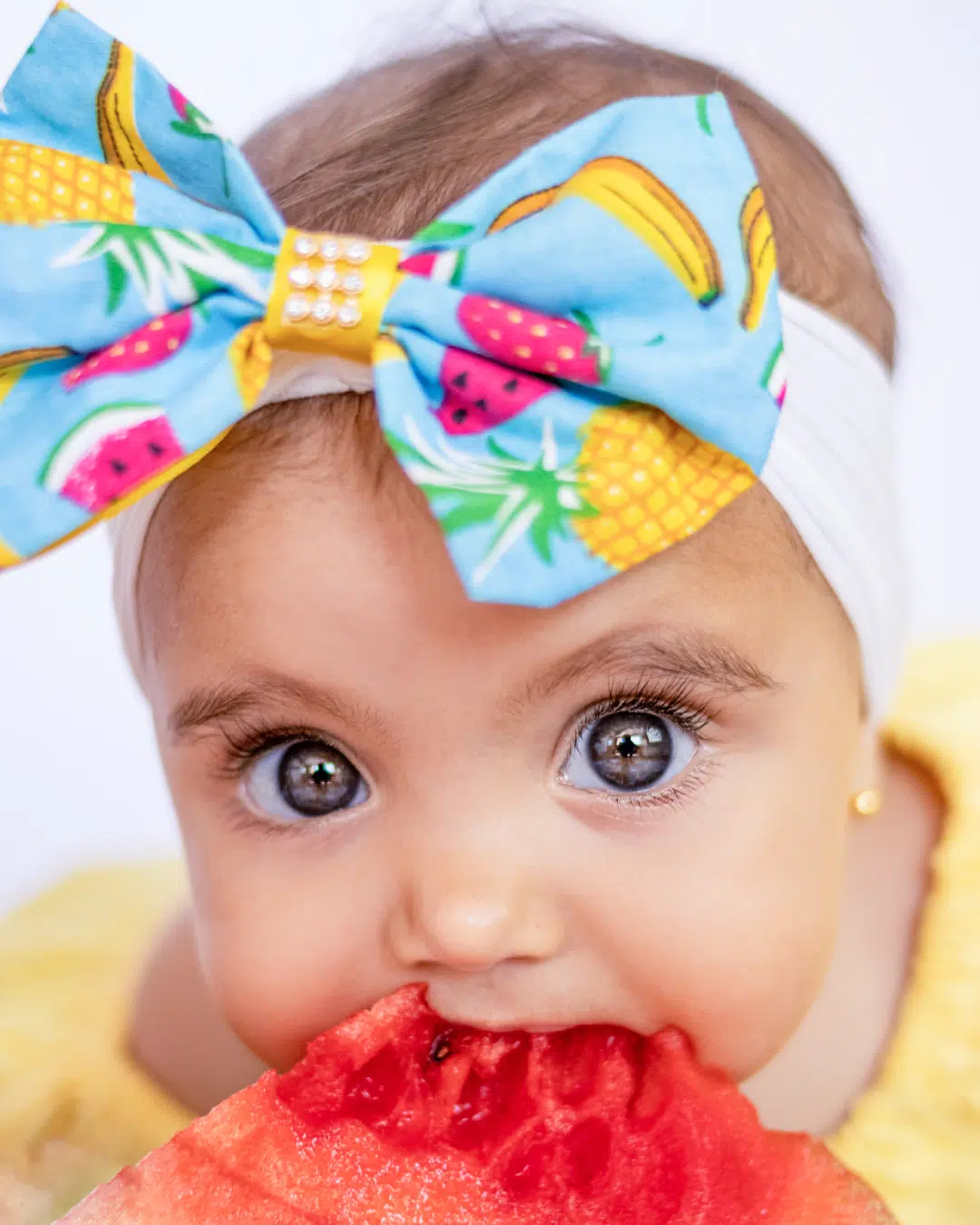 Retrato de bebê com laço colorido na cabeça, olhos brilhantes e segurando uma fatia de melancia, destacando o trabalho de pós-produção fotográfica de Carla Toscano Pós-Produção Fotográfica.